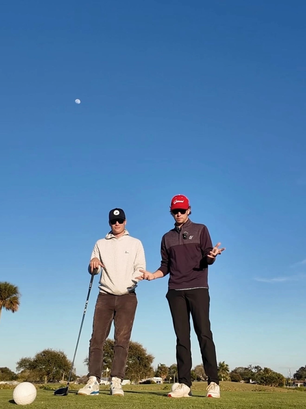 Kadin in a red cap shrugging on the tee box next to a friend, with a driver head lying on the grass