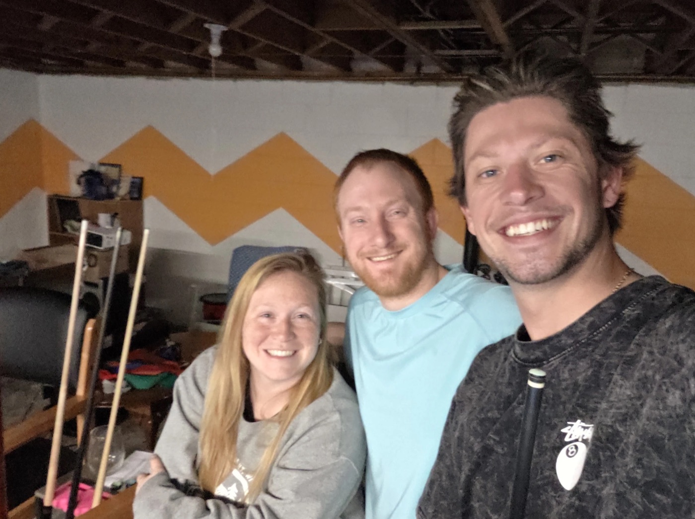 Kadin holding a pool cue in a basement selfie with two friends