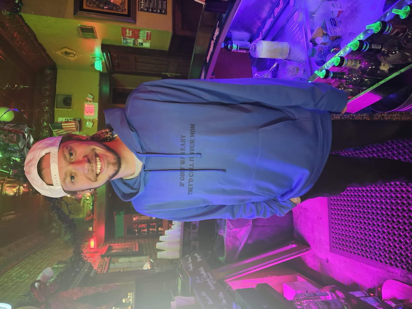 Kadin smiling behind a bar in a blue hoodie that reads 'If golf was easy they'd call it your mom'
