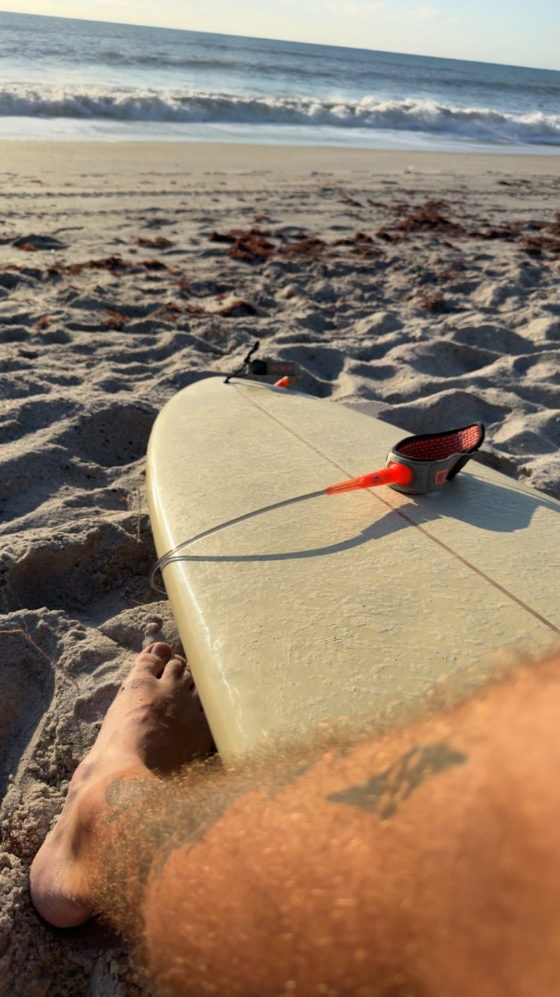 A surfboard resting on the sand with a foot in frame, ocean waves in the background