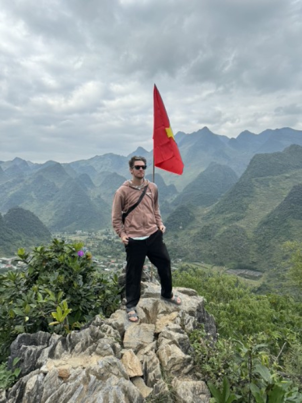 Kadin standing on a rocky peak in Vietnam beside a Vietnamese flag, green mountains behind him