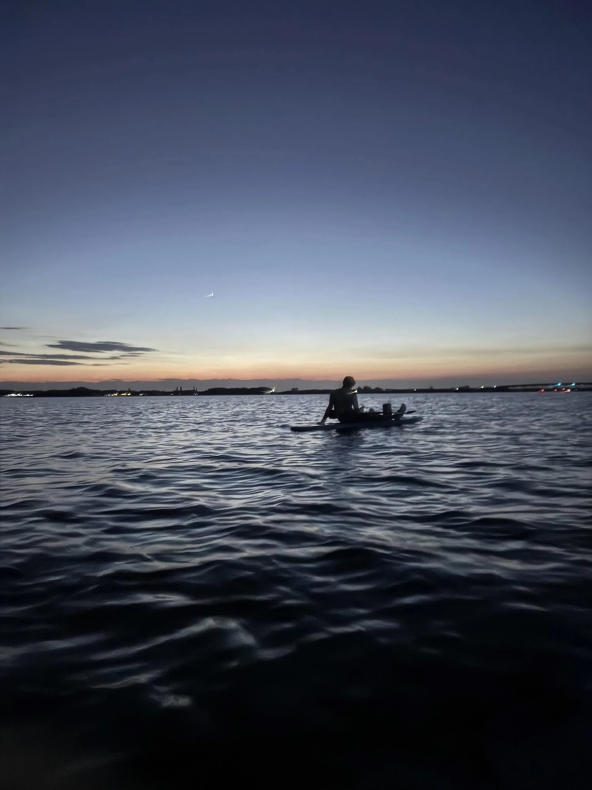 Kadin silhouetted on a paddleboard on calm water at dusk, sunset glowing on the horizon